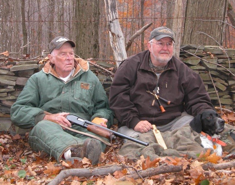 Two hunters sit against a stone wall in the fall turkey woods.