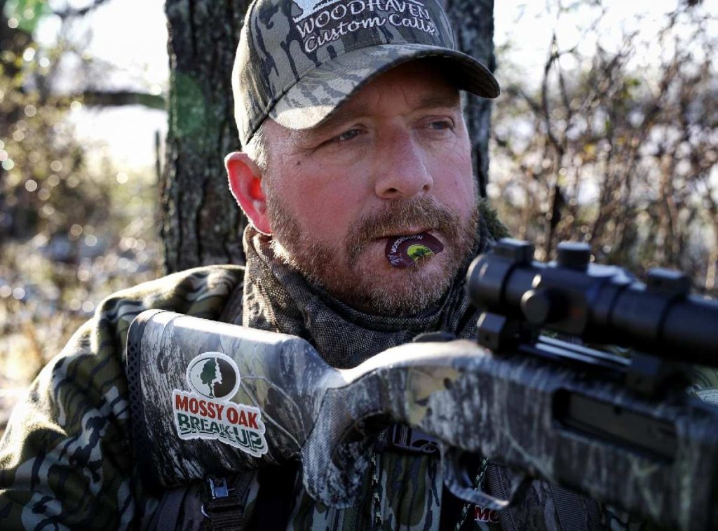 A hunter leans against a tree with a crossbow and a turkey call.