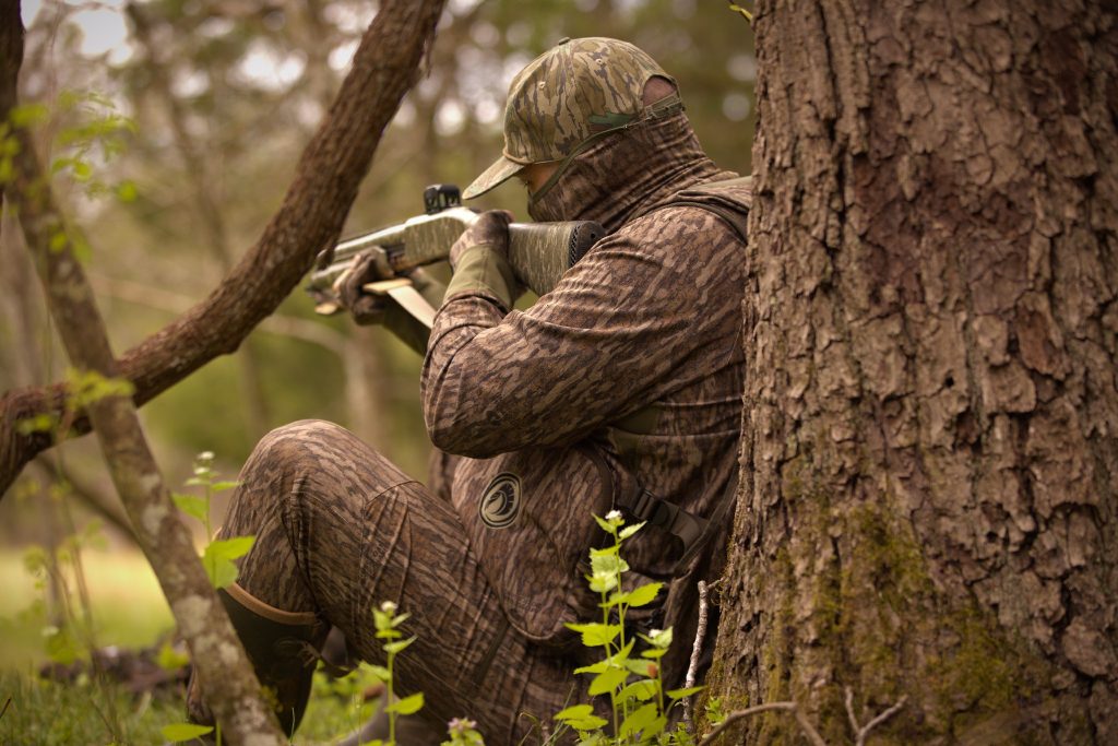 A hunter leans up against a tree and takes aim with a shotgun.
