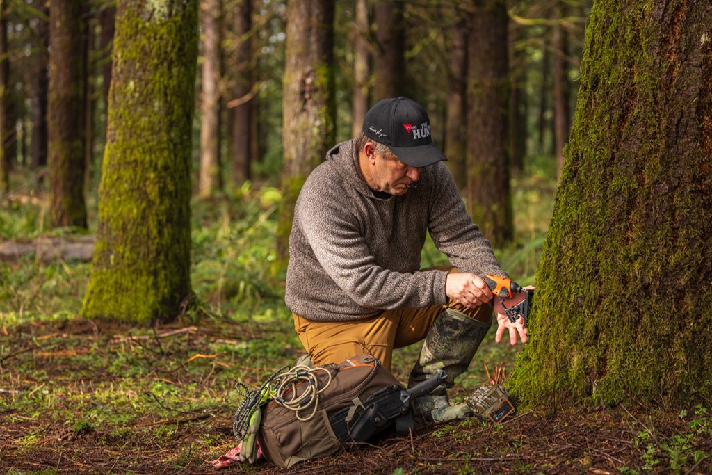 A hunter places a trail camera low on a mossy tree.