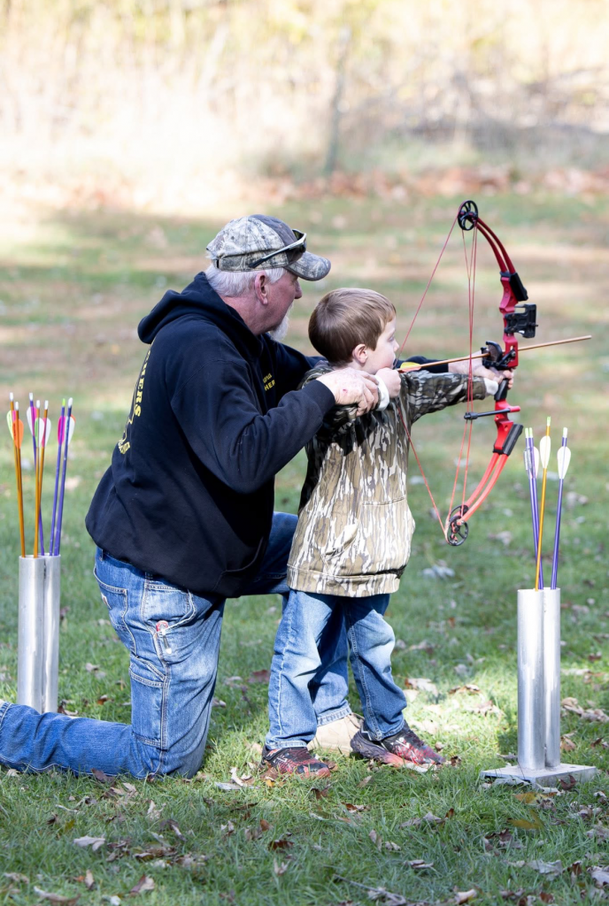 Young boy uses a compound bow with the help of a volunteer.